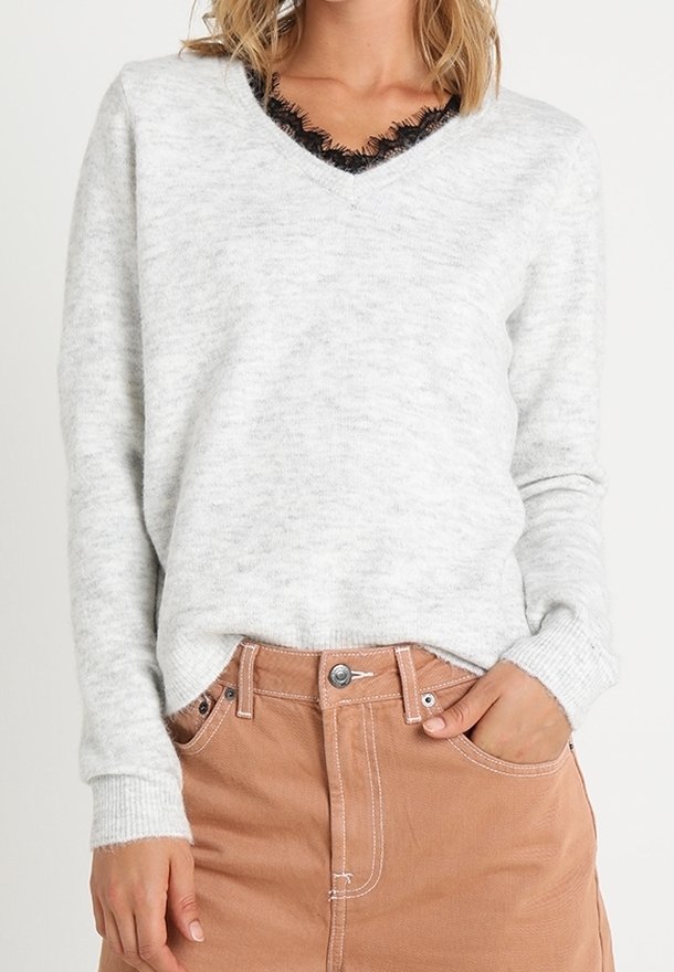 Femme portant un pull gris clair à col en V avec un détail en dentelle noire et une jupe taille haute beige avec des poches, main reposant dans la poche.
