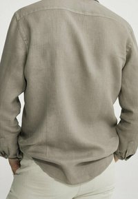 Lysebrunlangærmet skjorte lavet af blødt, tekstureret stof med en buet kant og manchetter med knapper. Bagfra vises et simpelt design.
