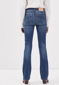 Jeans de denim azul de tiro alto con pierna acampanada, que cuentan con dos bolsillos traseros con costuras onduladas y una etiqueta de marca de cuero en la cintura.