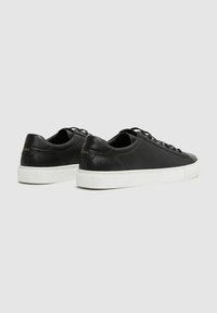Schwarze Ledersneaker mit runder Spitze, flacher weißer Sohle und Schnürung vorne. Die Rückseite zeigt den Markennamen in goldener Akzent auf der Ferse.