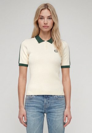 Femme blonde portant un polo en maille crème à manches courtes avec col et bords de manches vert foncé, associé à un jean bleu clair.