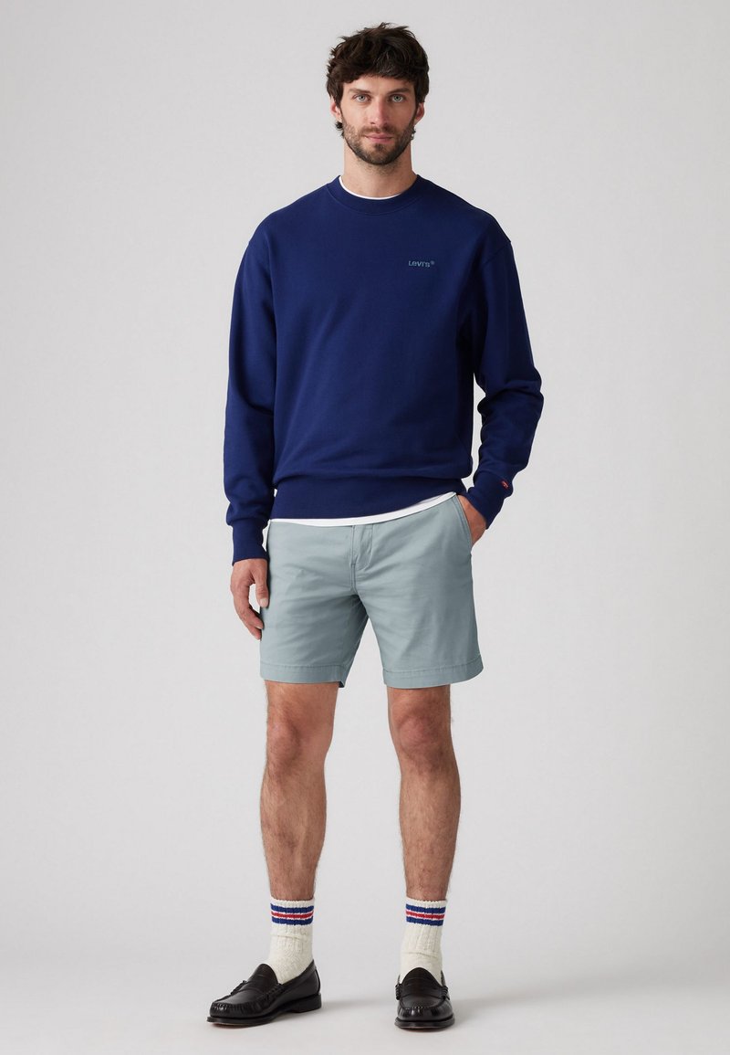 Sweat-shirt marine à col rond avec logo, associé à un short bleu clair. Des mocassins noirs et des chaussettes à motifs complètent la tenue.