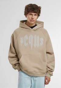 Felpa beige con tasca a marsupio, caratterizzata da audace testo grafico bianco sul davanti, realizzata in morbido materiale con vestibilità rilassata e polsini rigati.