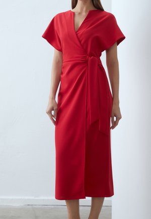 Woman wearing a red wrap dress with short sleeves and a tie detail at the waist, standing against a plain light background.