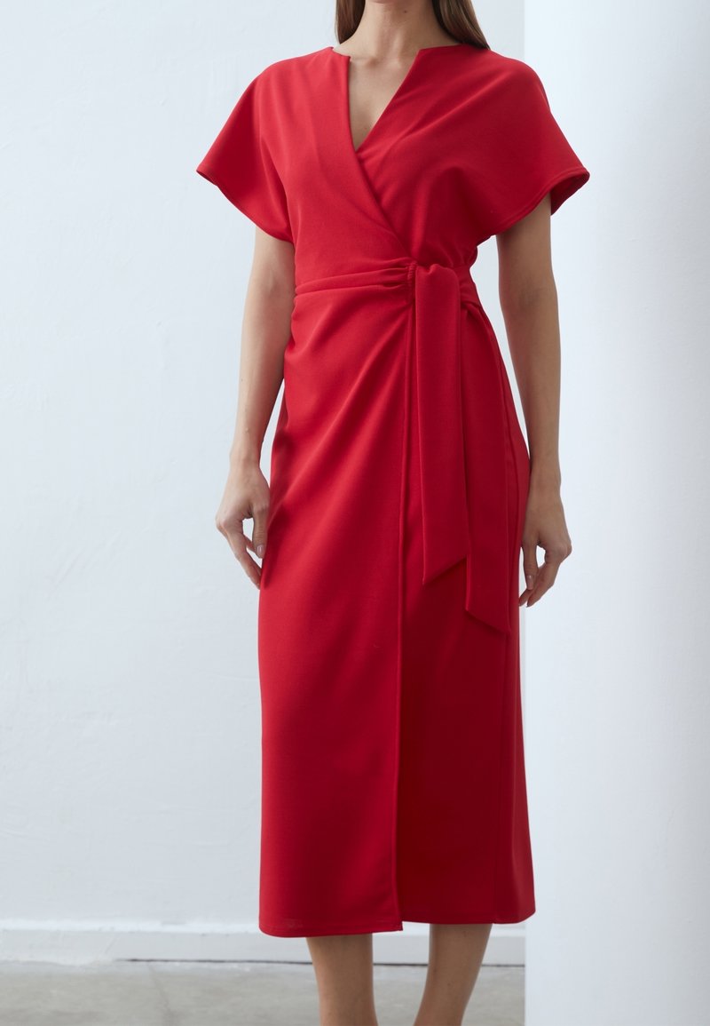 Femme portant une robe portefeuille rouge à manches courtes avec un détail noué à la taille, debout devant un fond clair uni.