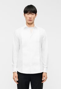 Λευκό πουκάμισο με κάθετο πλισέ σχέδιο, μακριά μανίκια, στενή γιακά και ίσια τελείωμα· λείο ύφασμα, μινιμαλιστικό στυλ.