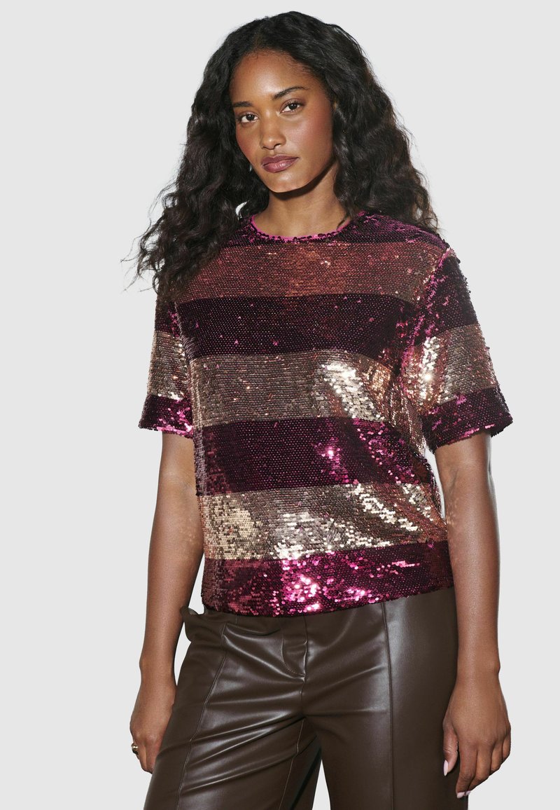 Paljettopp med horisontella ränder i djupröd, rosa och guld. Kort ärm, rund halsringning och en figurnära design. Kombineras med bruna byxor.