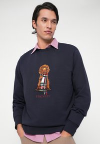 Mladenič v mornarsko modrem puloverju s grafiko psa in zasnovo tartan rute, oblečenem čez roza srajco s ovratnikom, z rokama v žepih.