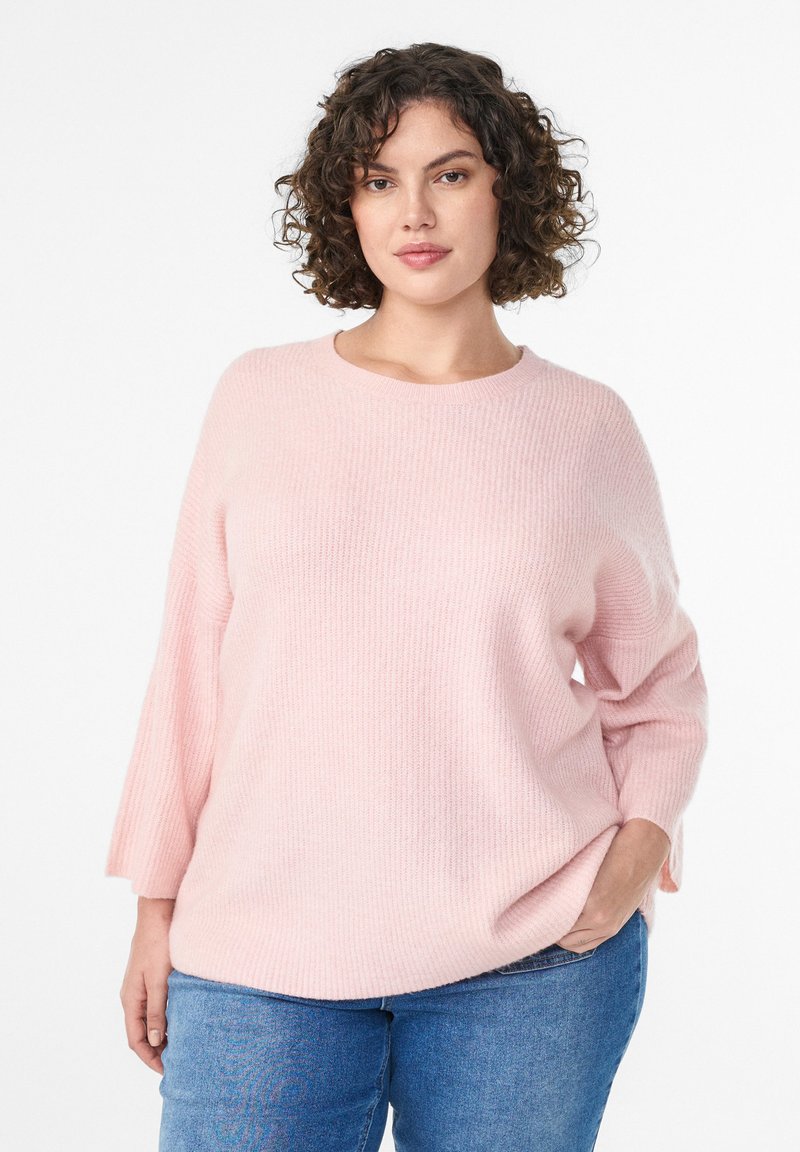 Light pink knitted sweater with wide sleeves and a round neckline. Worn over blue jeans, featuring a soft texture and relaxed fit.