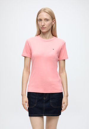 Femme aux cheveux blonds et raides, portant un t-shirt rose à manches courtes et une jupe en jean bleu foncé, debout devant un fond uni.