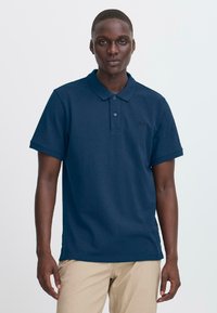 Mann iført en marineblå kortermet polo-skjorte og beige bukser, står mot en ensfarget lys bakgrunn.