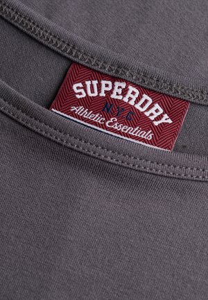 Zbliżenie na szarą tkaninę przy dekolcie z czerwoną metką Superdry NYC Athletic Essentials wszytą wewnątrz ubrania.