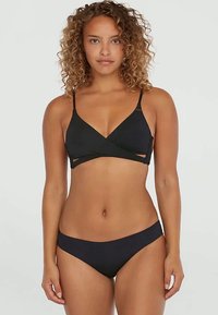 Nainen, jolla on kiharaiset hiukset, pukeutuneena mustaan kietaisumalliseen bikinitoppiin ja yhdisteisiin mustiin bikineihin, seisoo tasaisen taustan edessä.