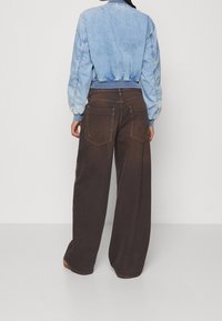 Džínová bunda ve světle modré barvě s žebrovaným límcem a manžetami, v kombinaci s tmavě hnědými kalhotami volného střihu, které mají zadní kapsy a švové detaily.