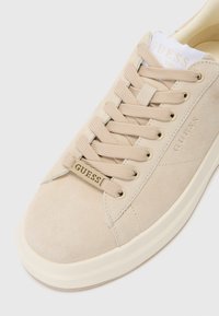 Beige Wildlederschuhe mit flachen Schnürsenkeln, einer abgerundeten Zehenpartie, Gummisohle und geprägt Logo an der Seite. Glatte Textur und minimalistisches Design.