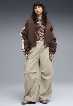 Femme portant une veste bomber marron sur un haut imprimé léopard, un pantalon beige à jambes larges, et des chaussures roses à pointes, sur un fond gris.