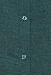 Teal skjorte med knappesluss, laget av teksturert stoff med et subtilt stripemønster, utstyrt med runde, mørke knapper langs frontplagget.
