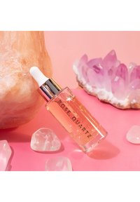 Szklana butelka z olejkiem różowego kwarcu, z białą zakraplaczem i złotym napisem na przezroczystej etykiecie, otoczona kryształami na różowym tle.