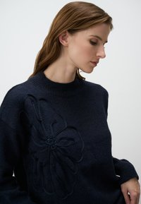 Un pull bleu marine se caractérise par un grand motif floral en relief sur le côté gauche, avec un col rond côtelé et un tissu texturé.