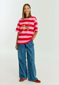 Červené a růžové pruhované tričko se žlutou kočkou s leopardími skvrnami, v kombinaci s širokými modrými džíny a červenými pantoflemi.