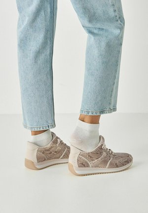 Světlé béžové tenisky s texturovaným vzorem připomínajícím hadí kůži, doplněné bílými ponožkami. Nošeny ke světle modrým džínům s krátkými nohavicemi.