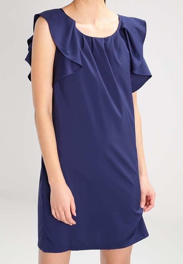 Femme portant une robe bleu marine sans manches avec des détails à volants sur les épaules et un col rond, debout devant un fond uni.