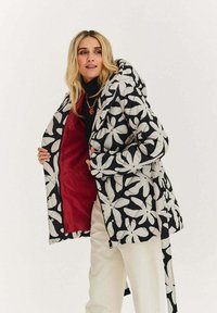 Černobílý prošívaný kabát s květinovým vzorem a červeným vnitřkem, který má zip a patentní knoflíky a měkký, strukturovaný materiál.