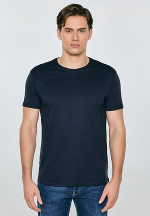 Jonge man met kort bruin haar, gekleed in een effen zwart T-shirt en blauwe spijkerbroek, staand tegen een lichtgrijze achtergrond.