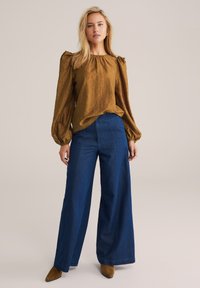 Braune Satinbluse mit Puffärmeln, kombiniert mit hochtaillierten, dunkelblauen, weit geschnittenen Jeans. Spitz zulaufende braune Stiefeletten runden den Look ab.