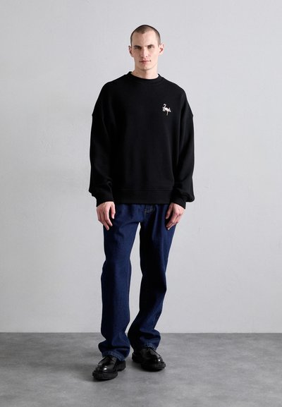 Sort sweatshirt med en afslappet pasform, ribbet halsudskæring og broderet design foran. Båret sammen med mørkeblå jeans og sorte sko.