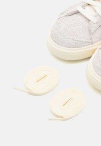 Sneaker in suede grigio con suole in gomma avorio. Due lacci piatti in cotone avorio posti accanto alle scarpe.