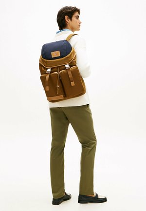 Giovane uomo con maglione bianco, pantaloni oliva, mocassini neri, che porta uno zainetto marrone e blu navy con tasche frontali, che guarda di lato.