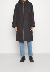 Manteau long noir à capuche avec fermeture éclair, grandes poches et manches ajustables, associé à un jean bleu clair et des bottines noires.