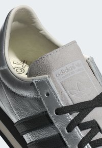 Metallic grey leather sneaker with black stripes, soft lining, textured tongue, and logo detail. Features round black laces.