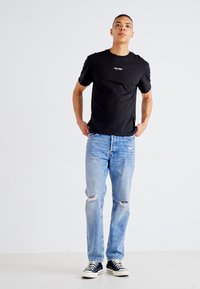 T-shirt noir en coton avec un col rond et un petit logo blanc, associé à un jean déchiré bleu clair et des baskets noir et blanc.
