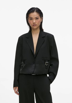 Kvinde med glat sort hår iført en sort skræddersyet blazer med spænde-detaljer og matchende sorte bukser, stående mod en hvid baggrund.