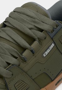 Gros plan sur une sneaker Globe vert olive avec des lacets épais, une languette en mesh, des détails cousus et une semelle en caoutchouc marron.