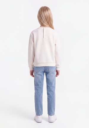 Barn med langt blondt hår iført en lysebeige langærmet top, lyseblå jeans og hvide sneakers, set bagfra.