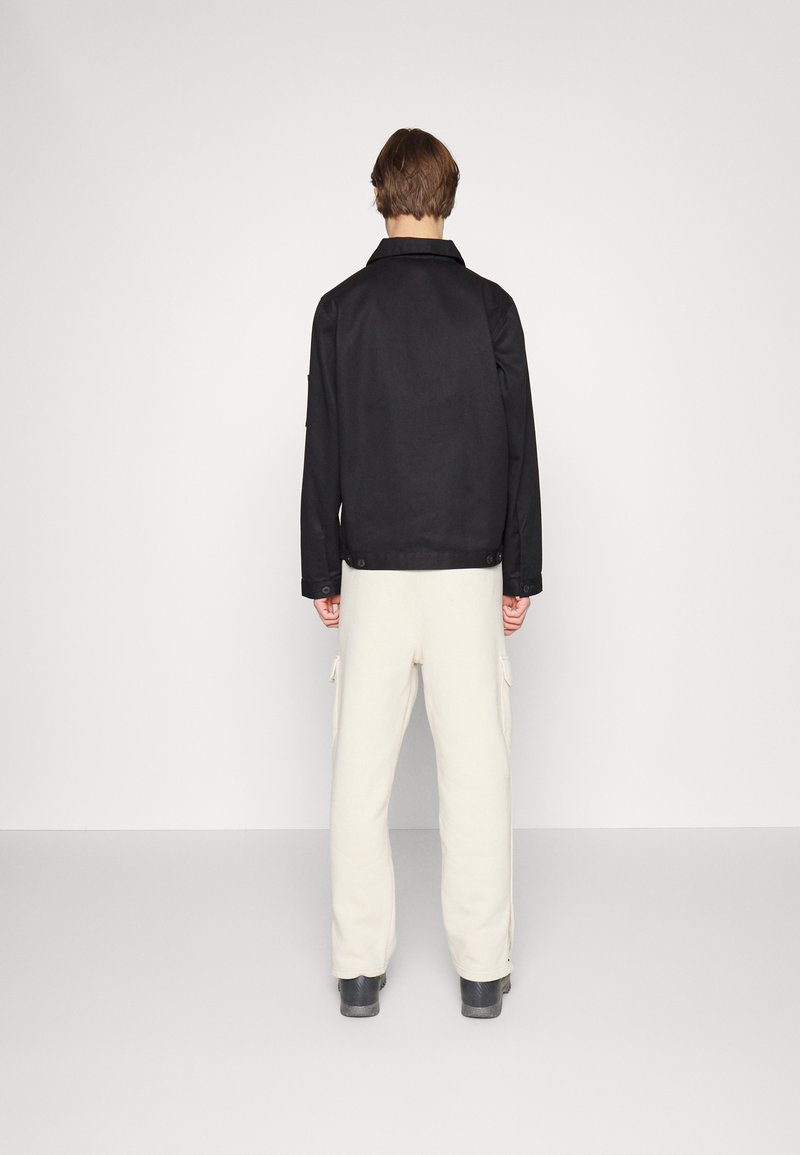 Giacca nera aderente con colletto, texture liscia e polsini abbottonati, abbinata a pantaloni cargo color crema e calzature nere, vista da dietro.