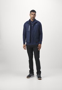 Giacca leggera blu navy con cappuccio, tasca sul petto con zip, abbinata a una polo navy e pantaloni neri. Scarpe da ginnastica completano il look.