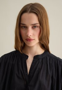 Schwarze, knöpfbare Bluse aus leichtem Stoff mit einem plissierten, hohen Kragen und Frontblende. Weiche Textur und lockere Passform.