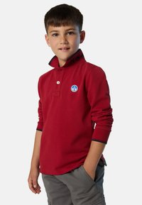 Polo a maniche lunghe rosso con patta a bottoni, colletto blu navy a contrasto e una toppa rotonda blu sul petto.