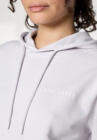 Svetlošedá mikina s kapucňou vyrobená z mäkkej tkaniny, s prednou vreckom, kapucňou so šnúrkou a bielym potlačeným logom "ATHLEISURE". Textúrovaný povrch, voľný strih.