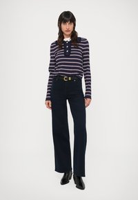 Marineblauw gestreept longsleeve shirt met een ronde hals, drie knopen en een donkere high-waisted jeans. Model draagt zwarte laarzen met hak.