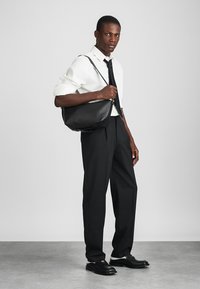 Fekete bőr messenger táska ívelt dizájnnal, állítható pánttal és sima textúrával, amelyhez egy testhez álló fehér ing és fekete nadrág párosul.