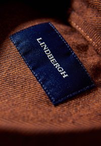 Granatowa tkanina z nadrukiem "LINDBERGH" w kolorze białym, wszyta na teksturowanym brązowym materiale. Krawędzie etykiety są lekko postrzępione, co podkreśla jej fakturę.