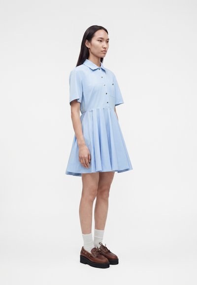 Γυναίκα που φορά ανοιχτό μπλε φόρεμα με κοντά μανίκια και γιακά, με χρυσά κουμπιά, λευκές κάλτσες και καφέ παπούτσια με πλατφόρμα, στέκεται σε απλό φόντο.