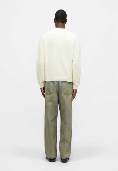 Férfi áll, háttal a kamerának, krémszínű pulóvert, laza, fakó zöldes-szürke farmert hátizsebekkel és fekete cipőt visel egy egyszínű háttéren.