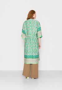 Vestido longo verde estampado com padrões florais, cintura ajustada e mangas largas. Combinado com calças largas castanhas e calçado leve.