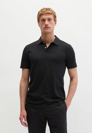 Młody mężczyzna o blond włosach, ubrany w dopasowaną czarną koszulkę polo z krótkim rękawem oraz czarne spodnie, stojący na jednolitym białym tle.